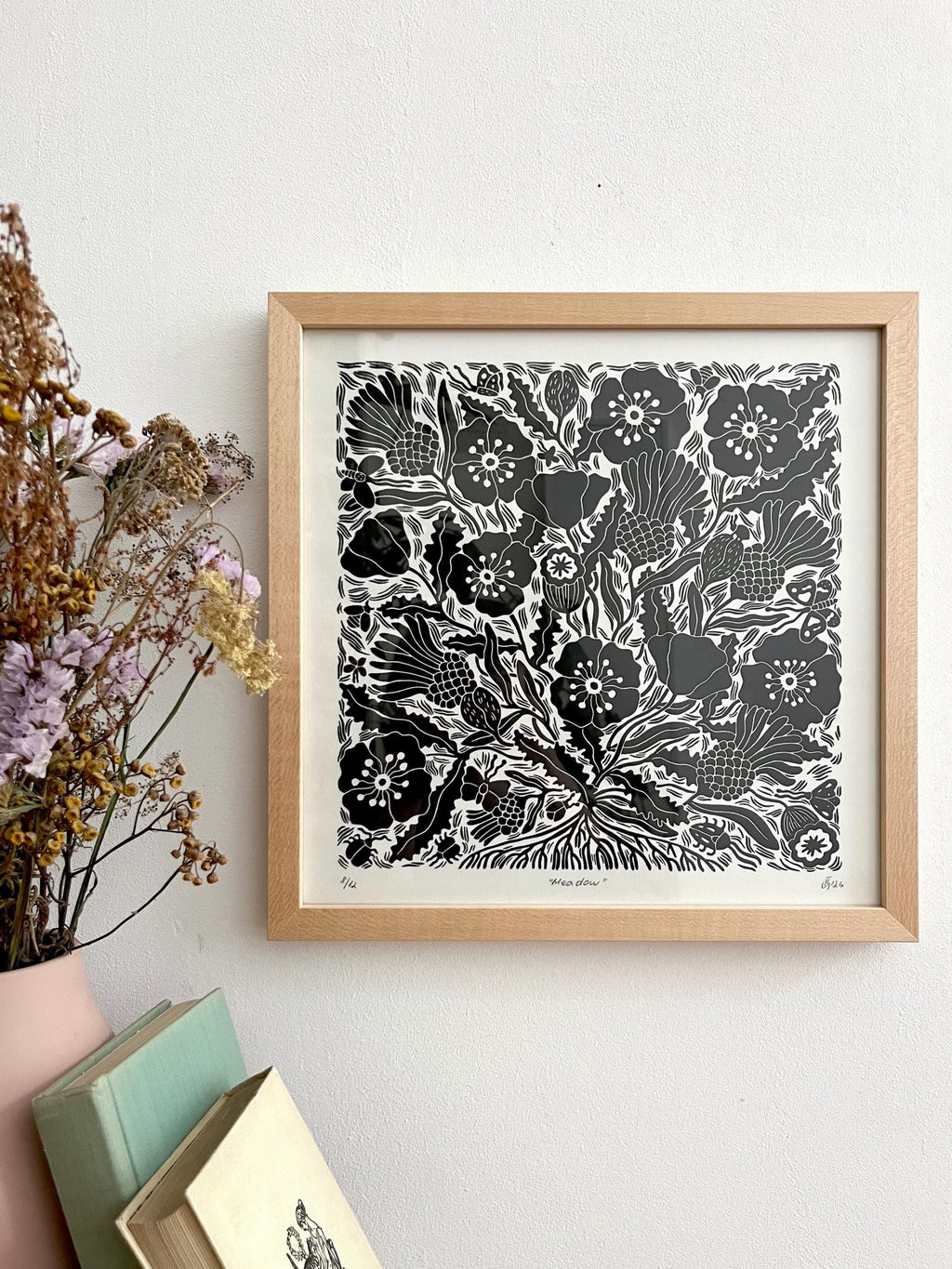A framed black and white floral linocut print with a light wood frame, hanging on a white wall next to dried flowers and stacked books.