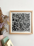 A framed black and white floral linocut print with a light wood frame, hanging on a white wall next to dried flowers and stacked books.