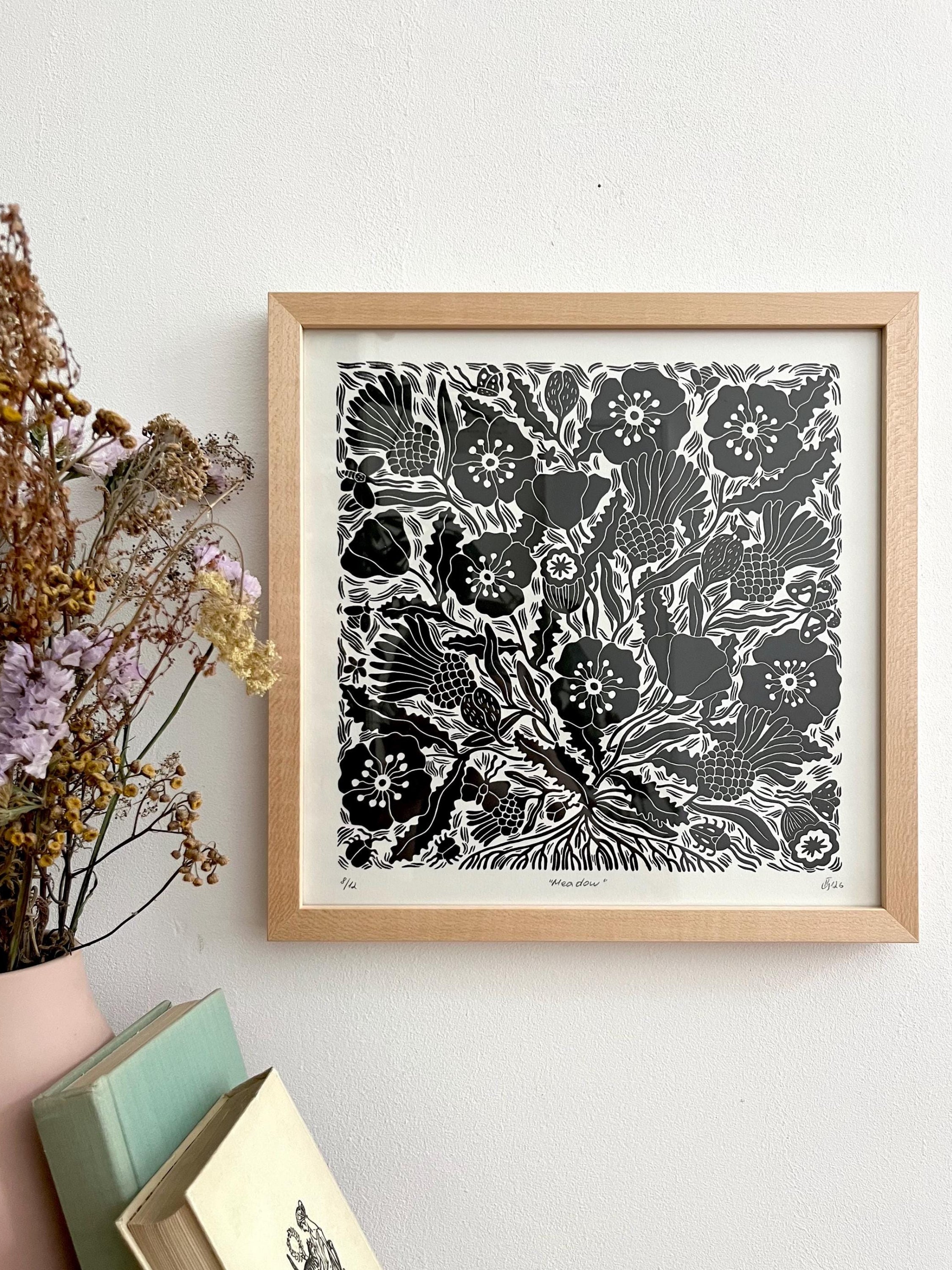 A framed black and white floral linocut print with a light wood frame, hanging on a white wall next to dried flowers and stacked books.