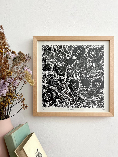 A framed black and white floral linocut print with a light wood frame, hanging on a white wall next to dried flowers and stacked books.